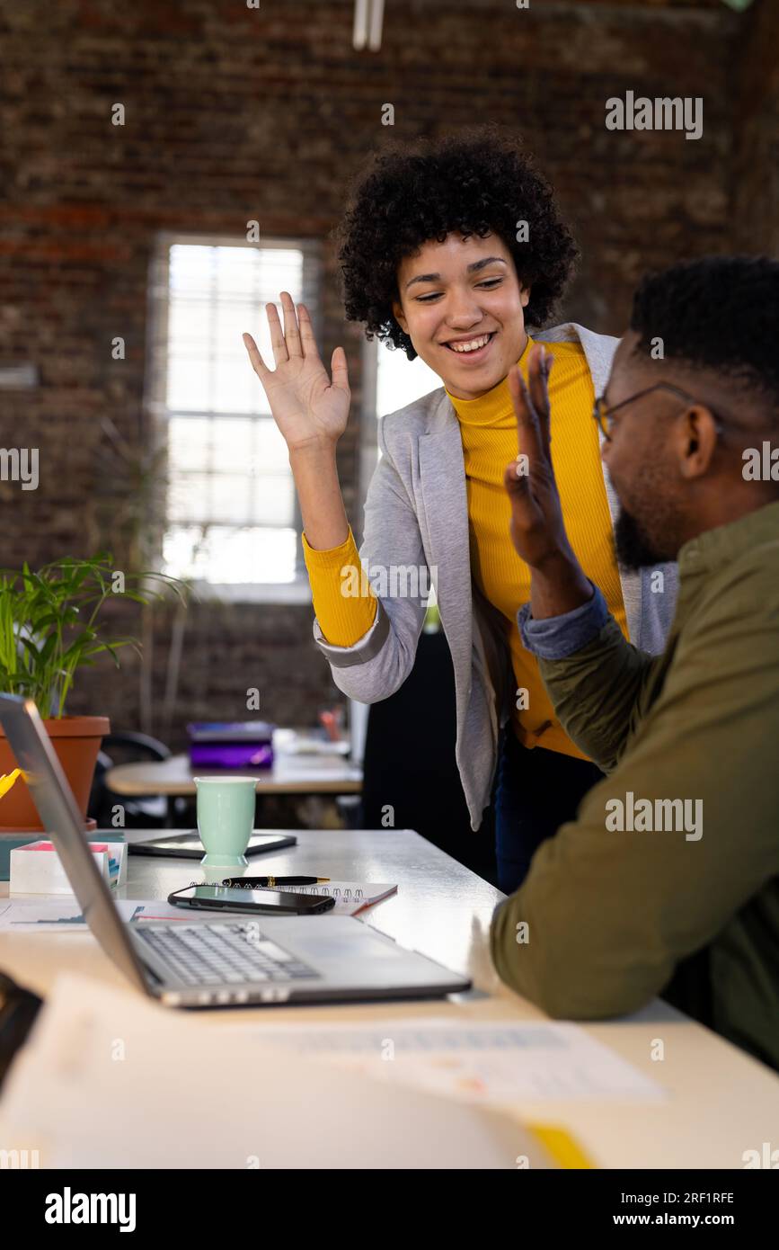 Heureux collègues masculins et féminins utilisant un ordinateur portable et high fiving au bureau dans un bureau occasionnel Banque D'Images