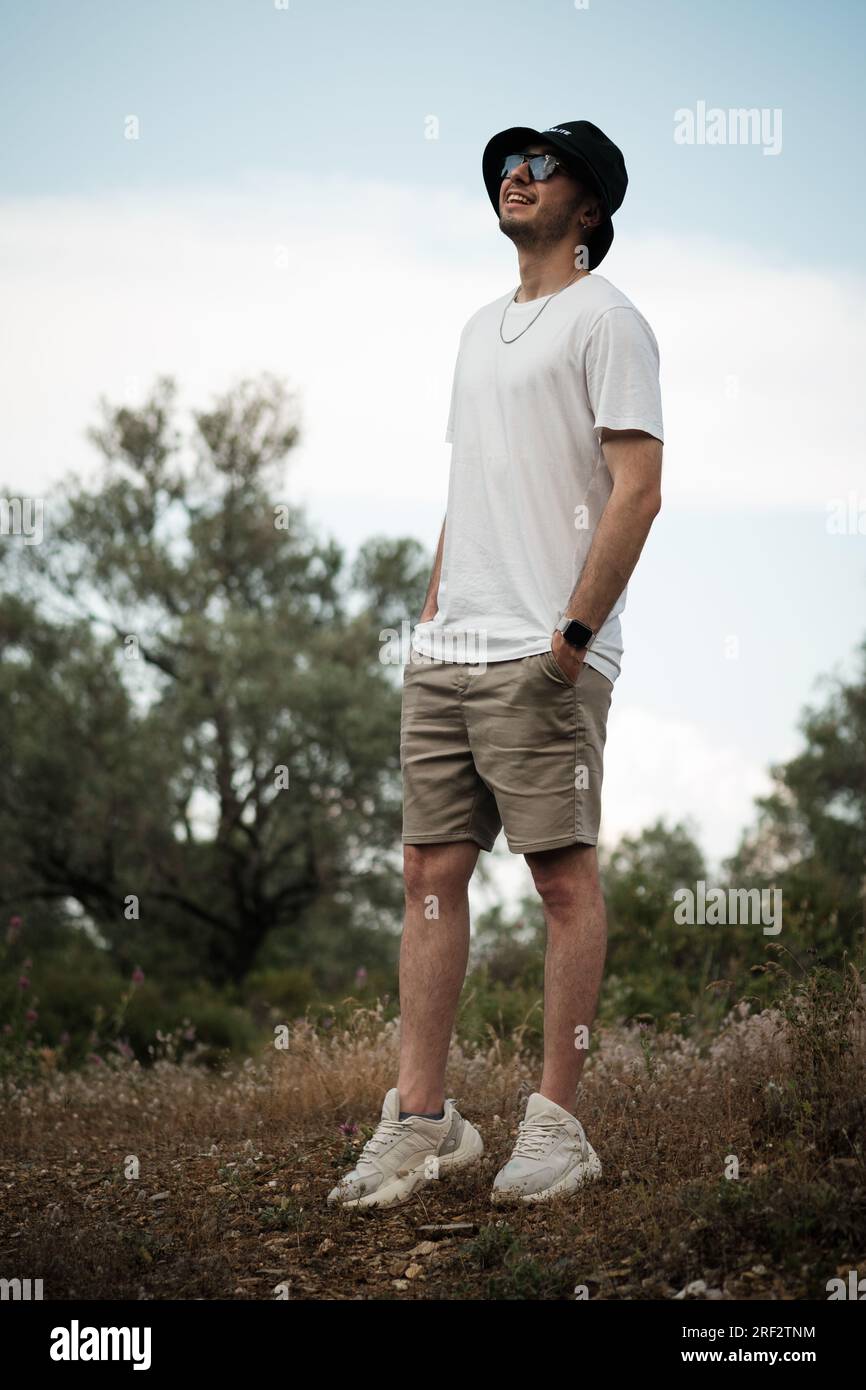 Un homme dans la vingtaine, riant ouvertement dans un t-shirt blanc, un short beige, des lunettes de soleil et une casquette de marin noire. Il est entouré par la beauté naturelle de Banque D'Images