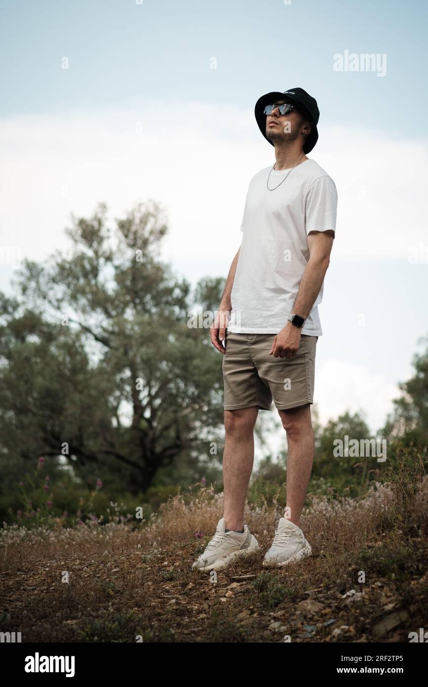 Un homme de 20 ans, vêtu d'un t-shirt blanc, d'un short beige, de lunettes de soleil et d'une casquette de marin noire, regardant sérieusement le ciel. Il est entouré par le Banque D'Images