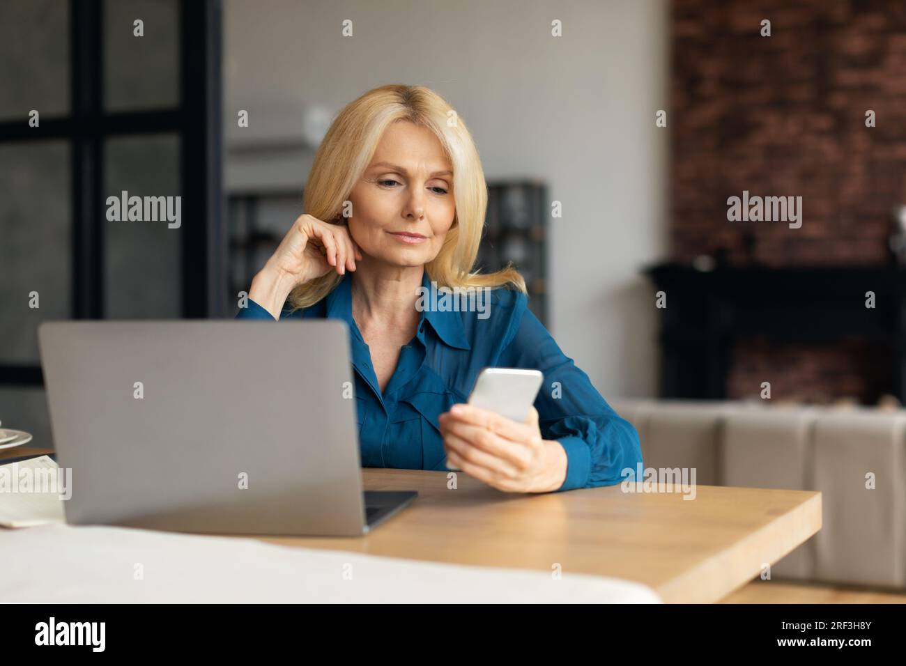 Femme d'affaires mûre focalisée travaillant de la maison, assise au bureau en face de l'ordinateur portable pc moderne et en utilisant le téléphone portable Banque D'Images