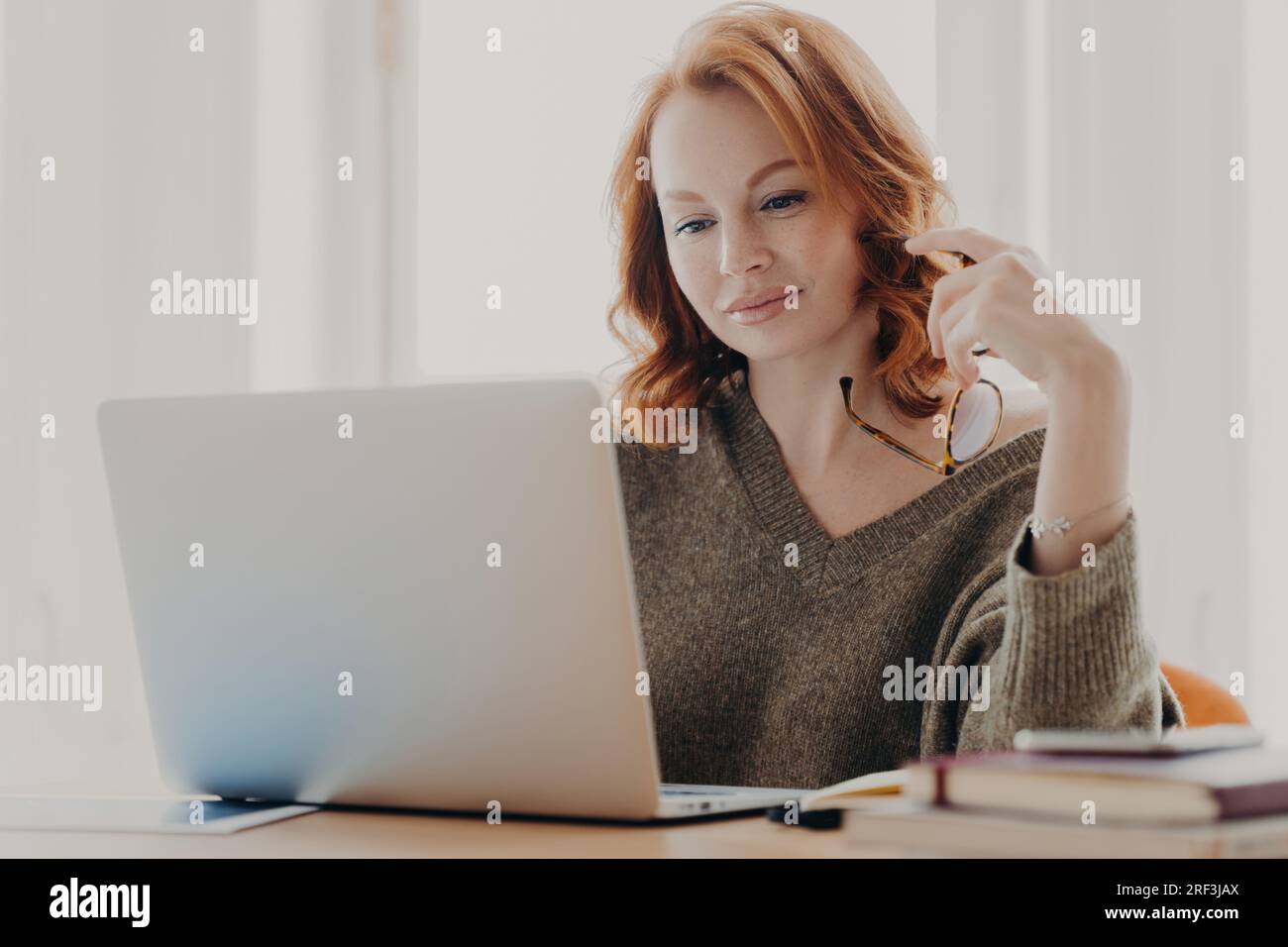 L'étudiante étudie attentivement, vérifie les données sur l'ordinateur portable, se prépare aux examens, utilise l'Internet sans fil, se concentre sur son espace de travail tout en tenant des lunettes pour Banque D'Images