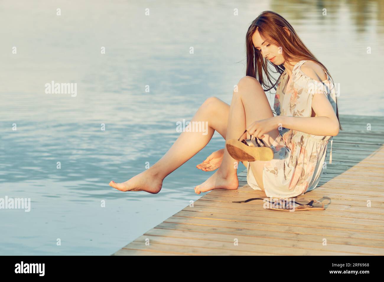 Jeune femme en robe de chambre s'assoit sur le bord de la jetée et enlève ses sandales pour tremper ses pieds dans l'eau Banque D'Images