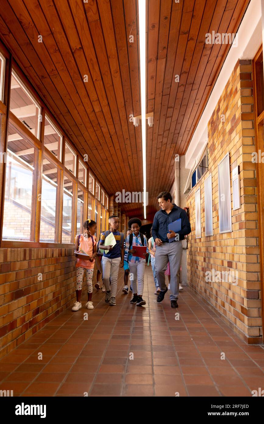 Enseignant masculin diversifié et enfants marchant ensemble dans le couloir à l'école primaire, espace de copie Banque D'Images