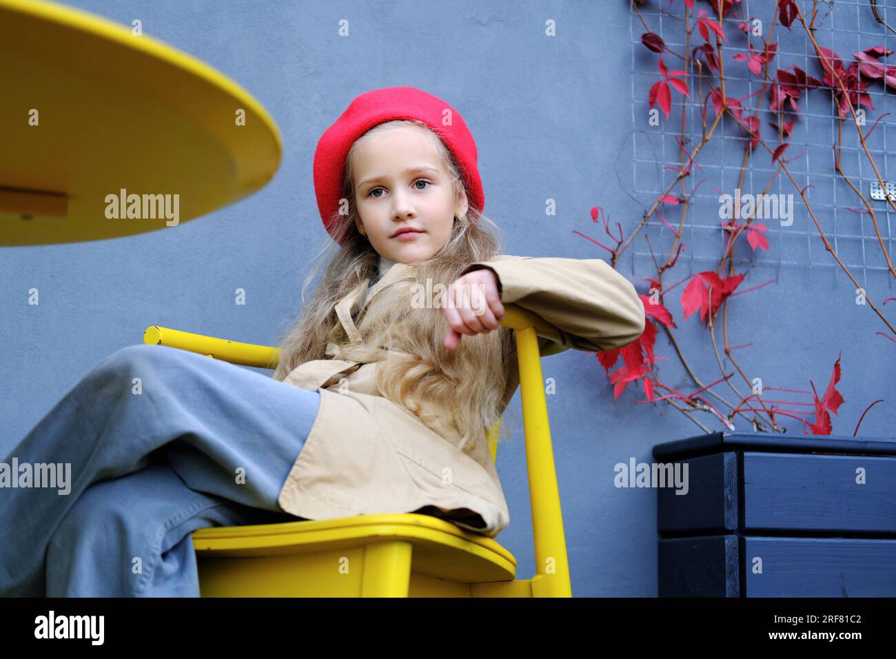 Belle petite fille dans un trench coat, béret rouge et jeans posant assis à la table d'un café de rue. Photo horizontale Banque D'Images