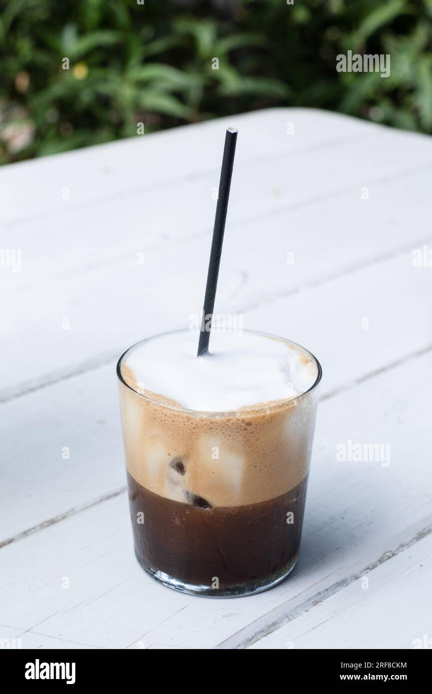 Cappucino fredo dans un gobelet sur une table en bois blanc, avec du café noir en bas, de la mousse de lait en haut et une paille de papier noire en haut. Banque D'Images