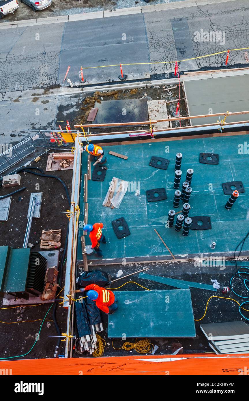 Travailleurs installant des matériaux de toiture sur le toit plat inférieur d'un bâtiment commercial en construction. Prise du toit supérieur du bâtiment. Banque D'Images