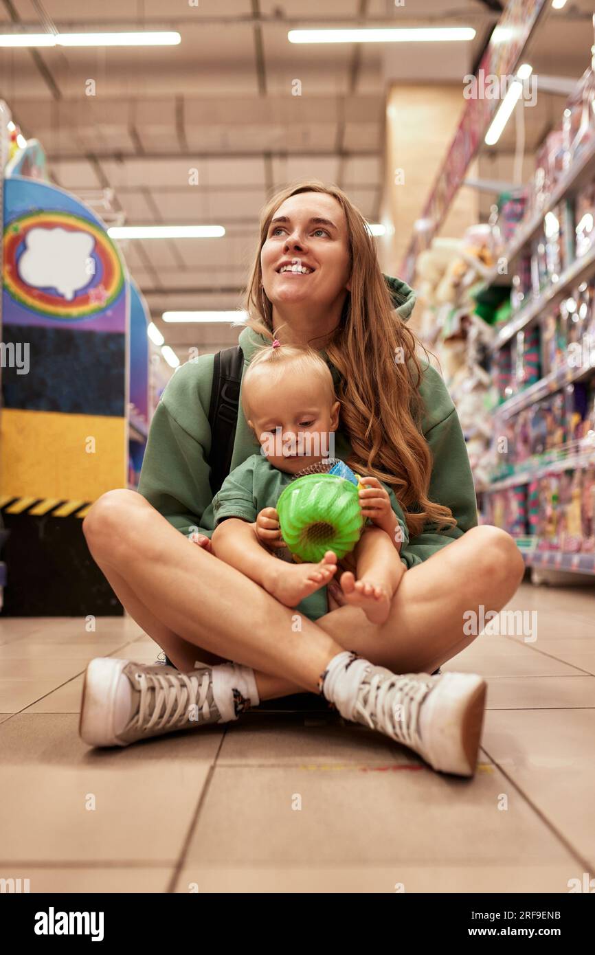 Heureuse jeune mère avec sa fille mignonne marchant autour du magasin de jouets dans le centre commercial. Maman et petite fille blonde choisissant des jouets et s'amusant. Amusant Banque D'Images