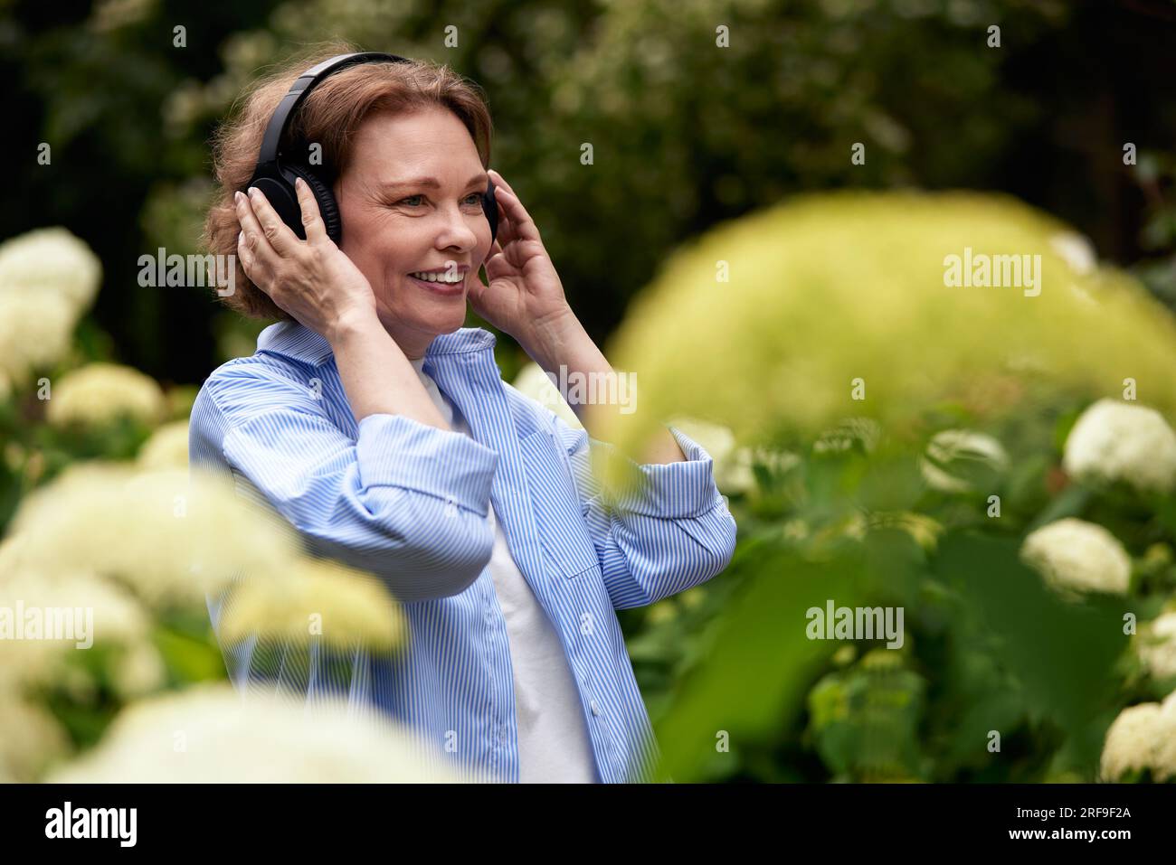 Portrait de femme mature attrayante dans le parc d'été avec des écouteurs sans fil. Dame senior appréciant la musique parmi les fleurs. Brunette d'âge moyen en bleu SH Banque D'Images