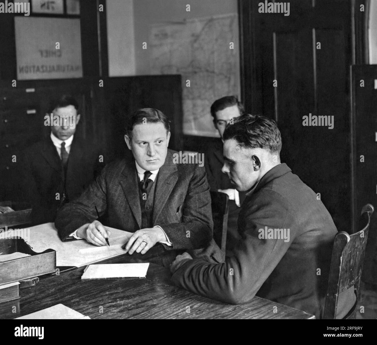 New York, New York : c. 1916 un immigrant est examiné au Naturalization Office à Ellis Island pour obtenir la citoyenneté américaine par un agent fédéral accompagné de deux témoins. Banque D'Images