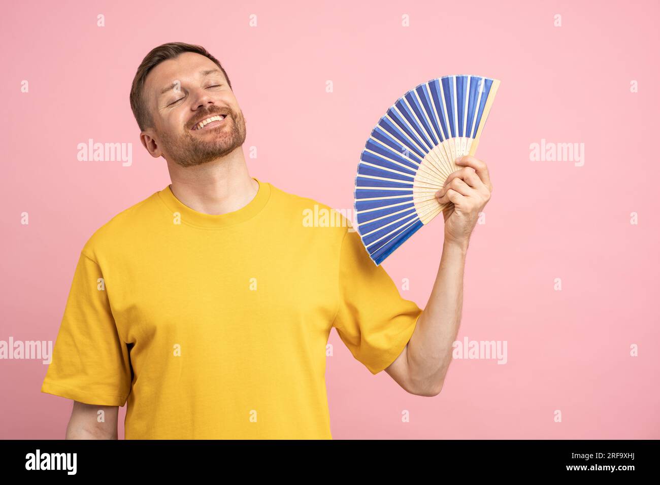 Homme surchauffé utilisant le ventilateur de papier souffrent de la chaleur, profitant de l'air frais isolé sur le mur rose studio Banque D'Images