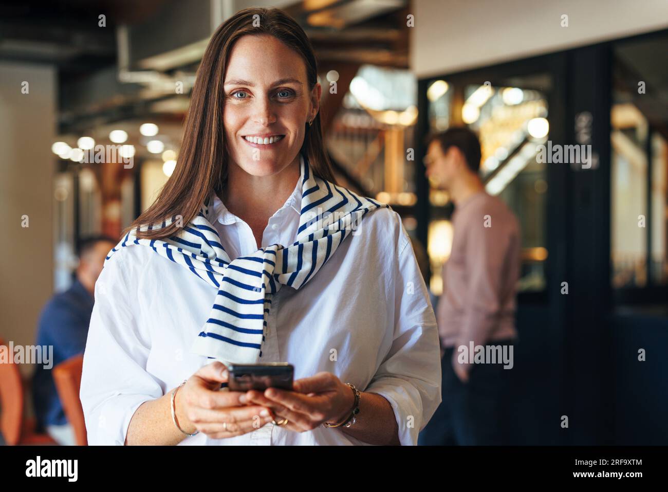 Femme d'affaires caucasienne tenant un téléphone portable et regardant la caméra. femme d'affaires debout dans un bureau avec ses collègues en arrière-plan. Banque D'Images
