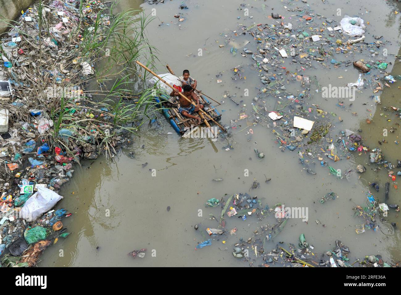 Un père et son fils rament sur un radeau fabriqué à la main à la recherche de déchets plastiques de recyclage dans un canal rempli de déchets plastiques de la zone Kazir Bazar de Sylhet. Ce canal est directement relié à la rivière Surma qui pollue la rivière Surma et remplit le lit de la rivière. Le polythène a accumulé du limon dans d'énormes tas, gênant la navigation et causant une pénurie d'eau utilisable de la rivière Surma au Bangladesh. Sylhet, Bangladesh. Banque D'Images