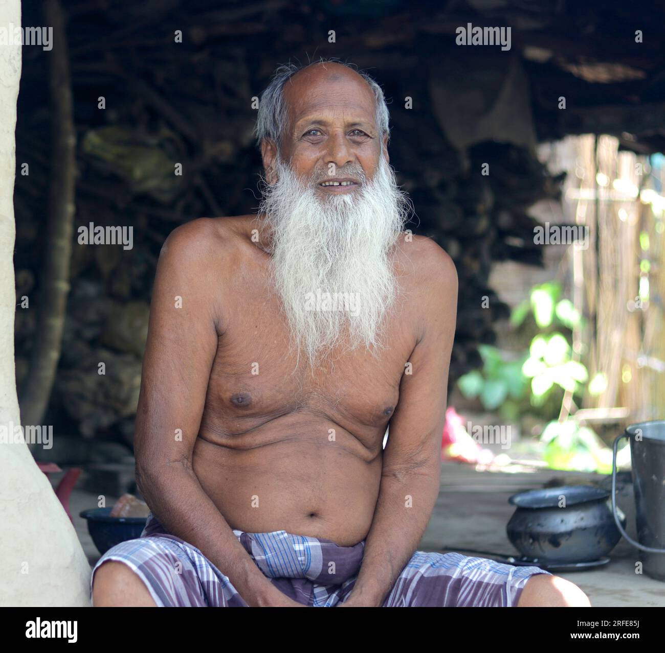 Un vieil homme pauvre avec une longue barbe blanche assis sur sa maison de boue de village. Banque D'Images