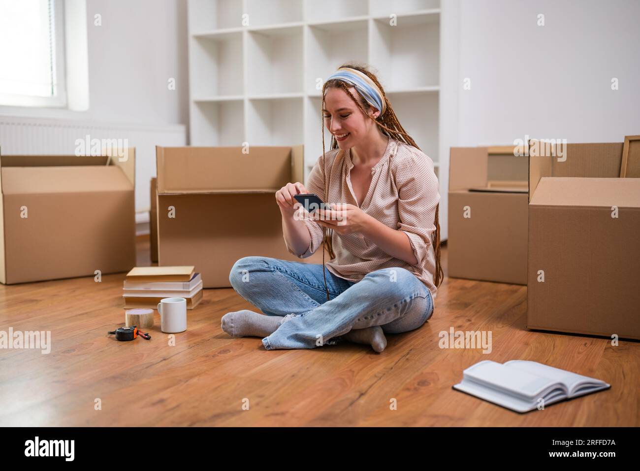 Femme moderne de gingembre avec des tresses utilisant le téléphone tout en emménageant dans son nouvel appartement. Banque D'Images