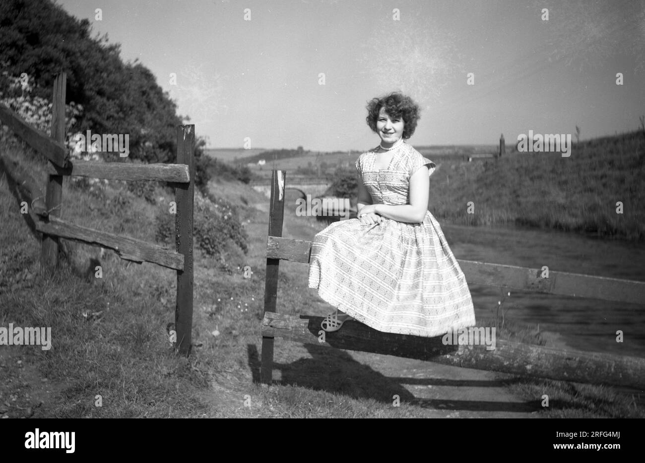 Années 1950, historique, une jeune femme en robe d'été de l'époque assise pour un sur une porte pour une photo sur un chemin de halage sur le canal Macclesfield, près de Congleton, Cheshire, Engand, Royaume-Uni. Banque D'Images