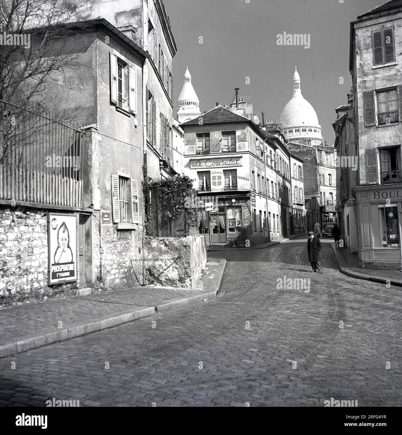 Années 1950, Paris, France, un homme marchant acorss une rue pavée dans le quartier Montmartre de la ville parisienne, célèbre pour ses artistes de rue et son église, le Sacré coeur. La photo montre les rue des Sales et le restaurant, 'le Consulatt' avec les dômes du Sacré-cœur en arrière-plan. Banque D'Images