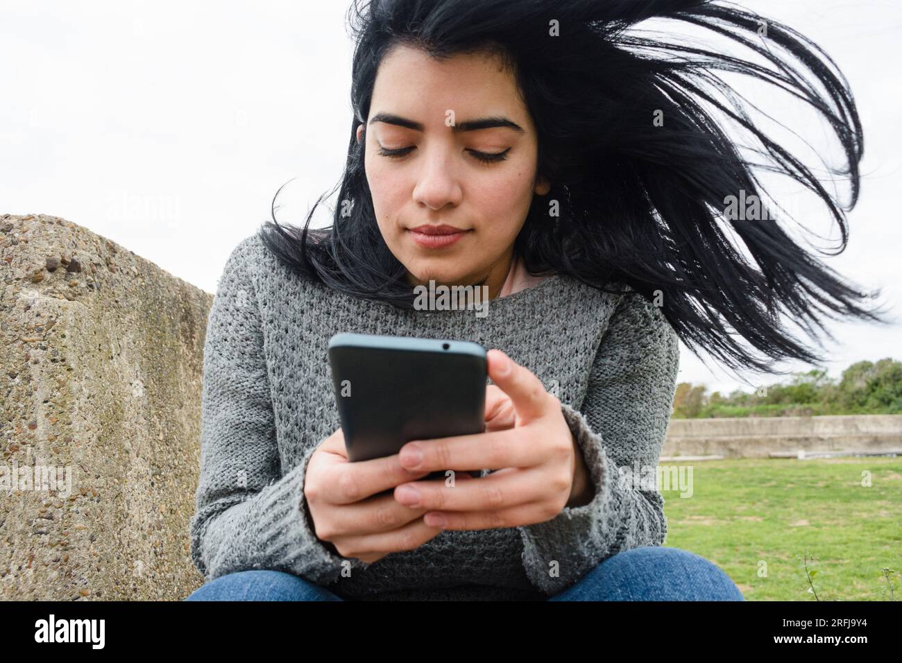 jeune femme latina vénézuélienne, utilisant le téléphone, lisant des messages, naviguant sur internet, tranquillement assise dehors par une journée nuageuse et froide, habillée Banque D'Images