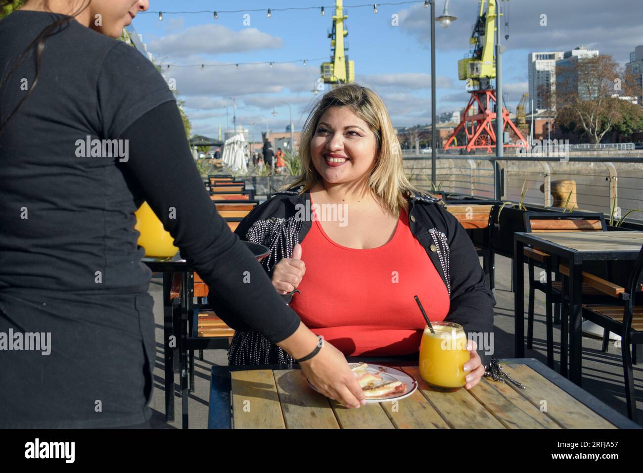 jeune femme latine blonde plus grande souriante très heureuse, assise à l'extérieur du restaurant au coucher du soleil, tandis que la serveuse lui sert un sandwich à l'orange Banque D'Images
