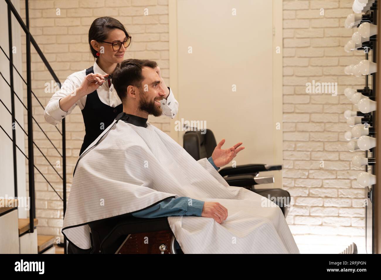 Client souriant assis dans un salon de coiffure dans une chaise de coiffure, une femme lui fait une coupe de cheveux avec des ciseaux Banque D'Images
