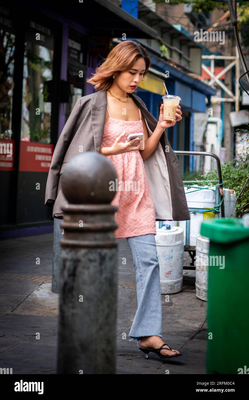 Une jeune fille thaïlandaise sirote son café et vérifie son téléphone en faisant son chemin vers Sala Daeng Rd. Bangkok Thaïlande. Banque D'Images