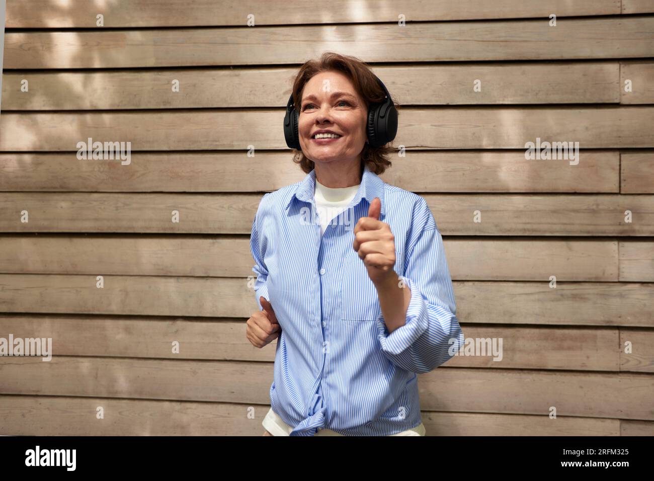 Portrait de femme mature attrayante avec des écouteurs sans fil sur fond de mur de chalet de parement. Brunette d'âge moyen dans une chemise bleue écoutant Banque D'Images