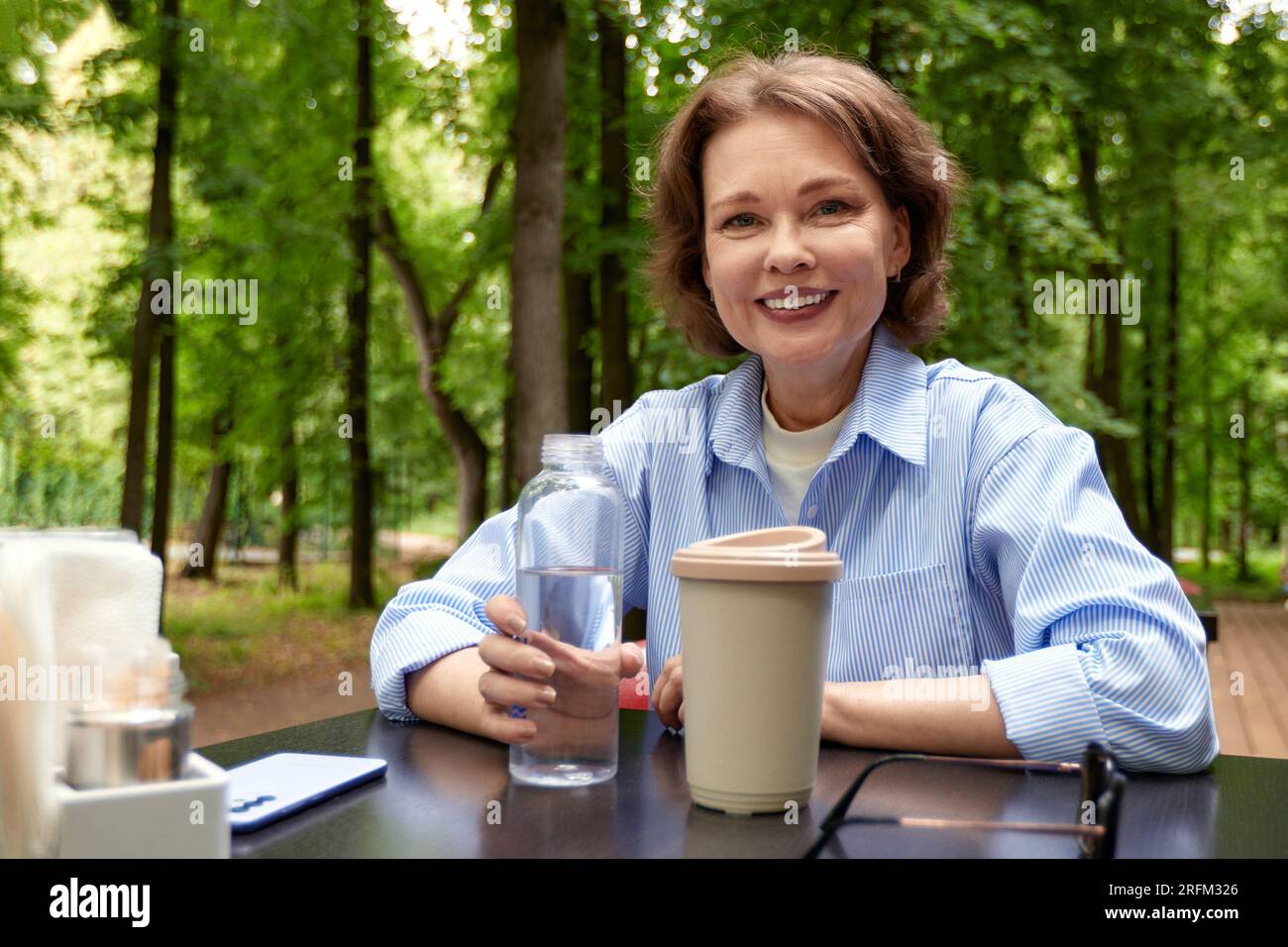 Adorable femme mature est assise à la table dans le café extérieur avec tasse à café en papier et bouteille d'eau. Dame senior appréciant la vie sur la terrasse d'été. Milieu Banque D'Images