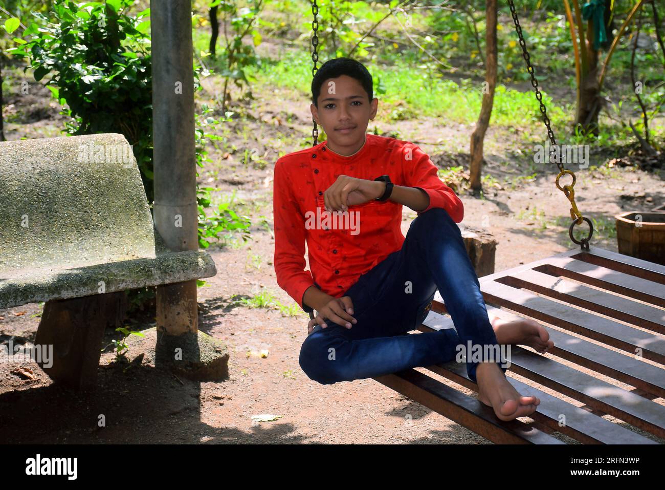 Garçon indien en chemise rouge et jeans bleu est assis dans une balançoire en fer. fond de jardin Banque D'Images