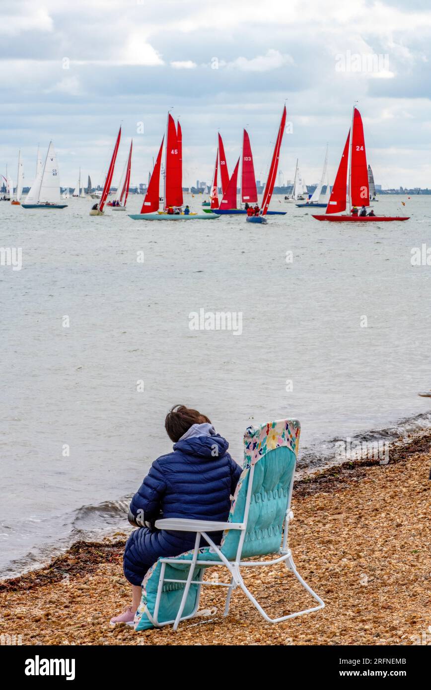 femme plus âgée assise dans une chaise pliante sur une plage de galets au bord de la mer regardant les yachts pendant la régate annuelle cowes week sur l'île de wight Banque D'Images