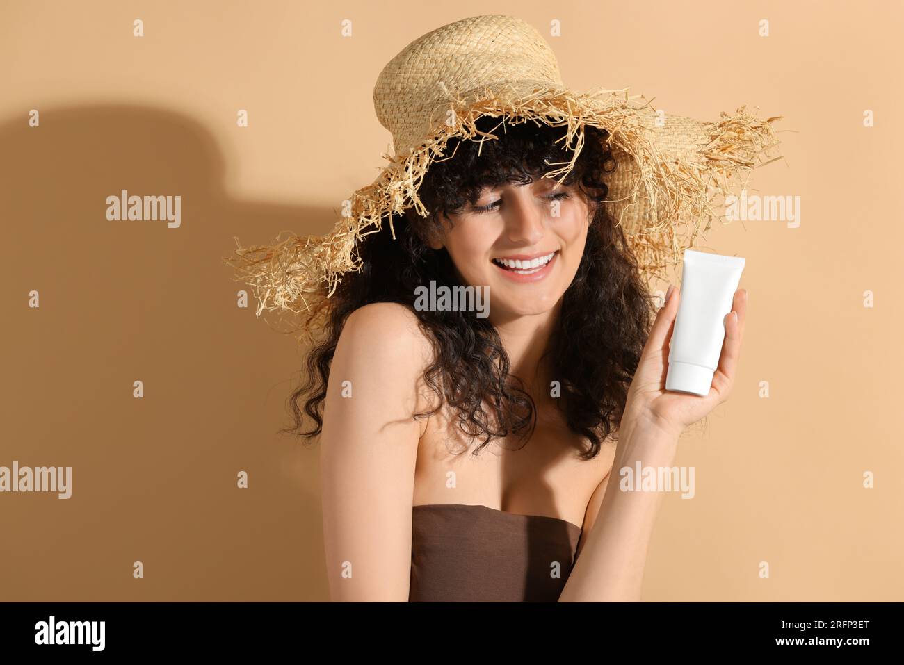 Belle femme heureuse dans le chapeau de paille tenant tube de crème de protection solaire sur fond beige Banque D'Images