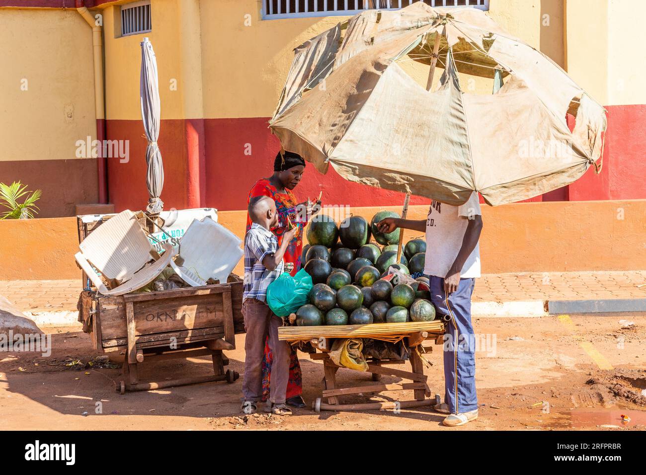 Abritée sous un parasol, une vendeuse de pastèques négocie avec une femme et son fils. Jinja, Ouganda. Banque D'Images