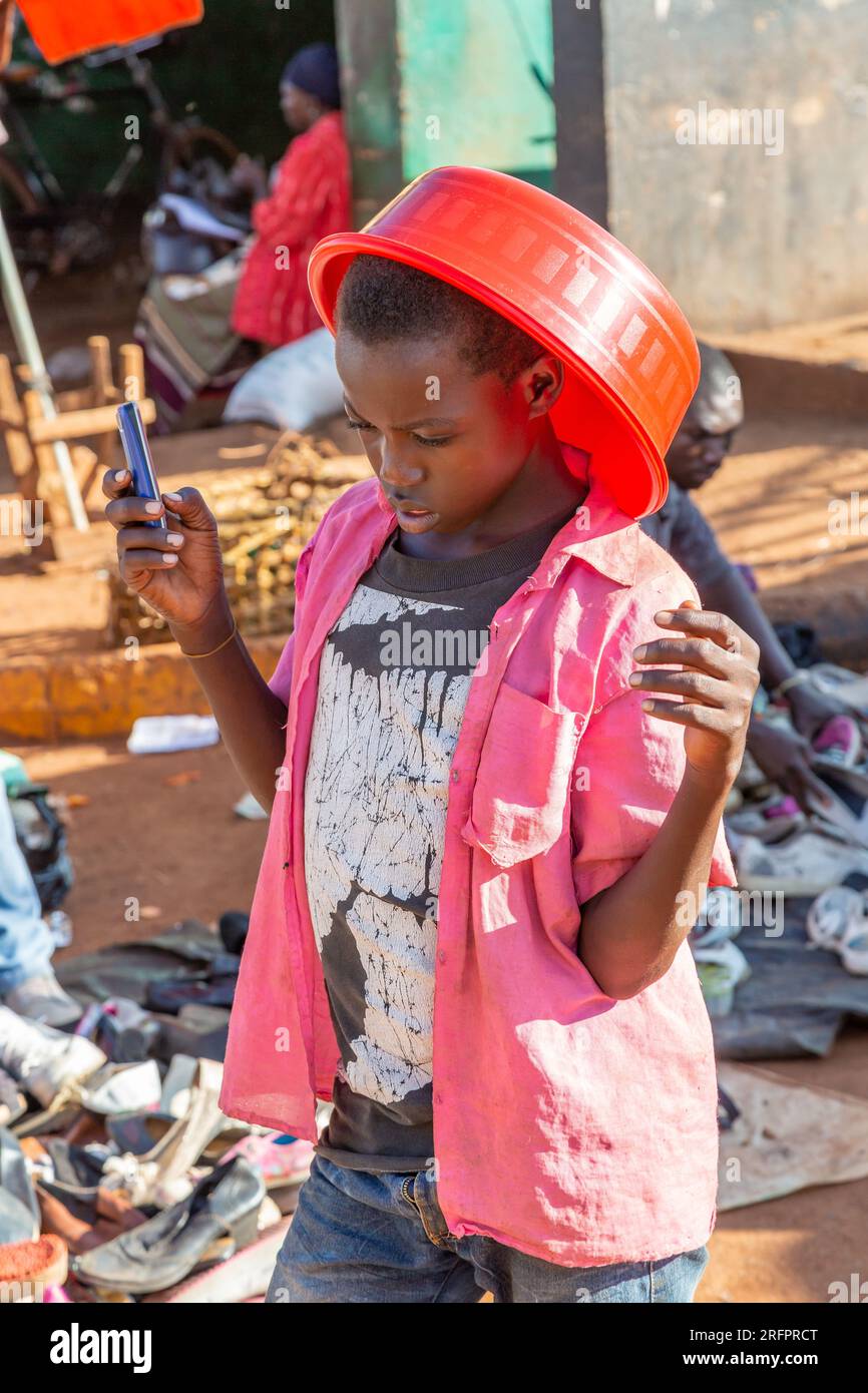 Au marché de Jinja : jeune garçon portant un bol en plastique rouge, téléphone portable à la main. Banque D'Images