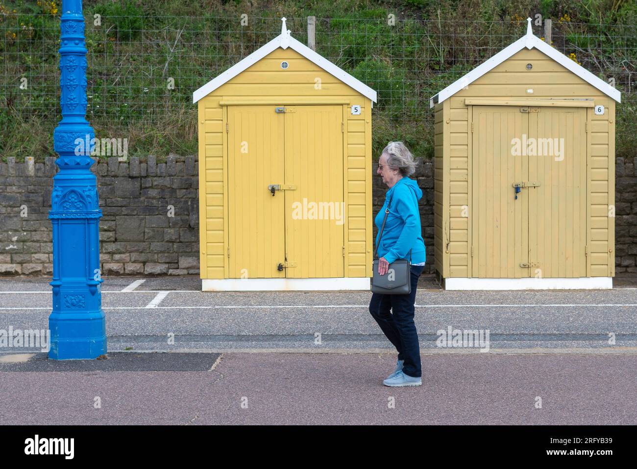 Femme portant un manteau bleu marchant devant des cabanes de plage jaunes, Bournemouth, Dorset, Angleterre, Royaume-Uni, 2 août 2023, Météo. Sauvage et venteux avec de fortes averses. Banque D'Images