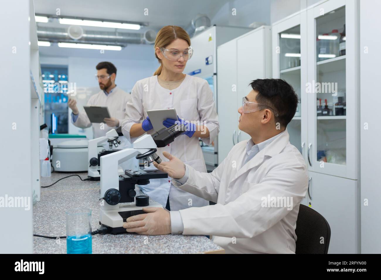 Travail dans le laboratoire d'une équipe internationale de scientifiques. Un homme asiatique examine sous un microscope et discute avec une collègue féminine. Un troisième homme en arrière-plan fait le point sur les drogues. Banque D'Images