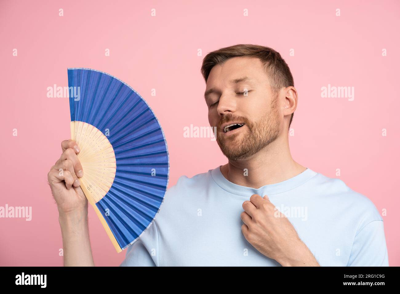 L'homme de surchauffe se sent soulagé le refroidissement tient des supports de ventilateur bleus sur fond rose de studio de mur Banque D'Images