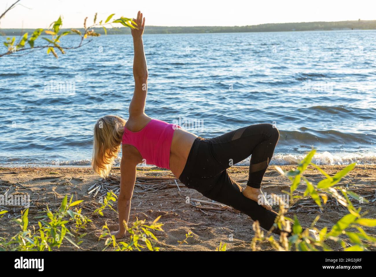 Une vue arrière d'une femme debout sur ses mains, étirant son bras vers le haut. Une femme pratiquant le yoga au bord du lac en été. Méditation. Sports d'été Banque D'Images