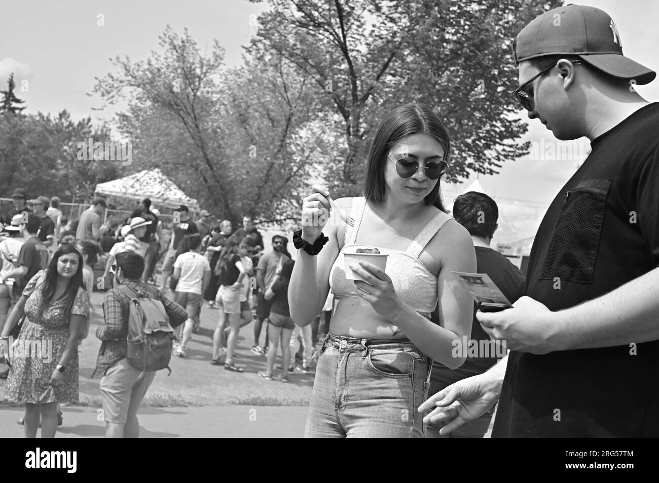 NOIR ET BLANC. 2023 Festival culinaire Taste of Calgary. Une jeune femme portant des lunettes de soleil en forme de cœur mange de la nourriture tout en parlant à son petit ami. Banque D'Images