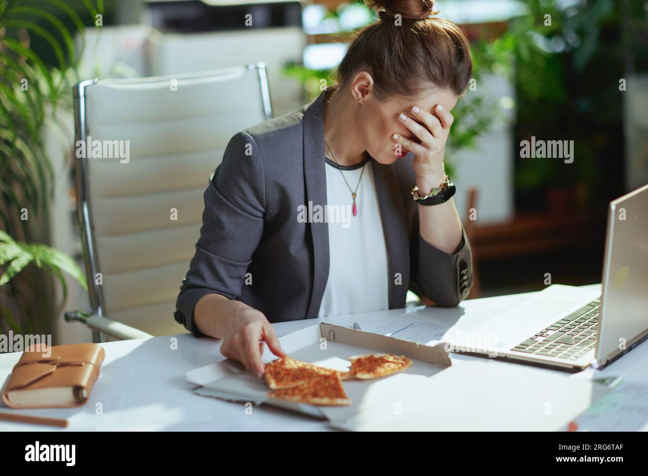 Lieu de travail durable. femme comptable moderne stressée dans un costume gris d'affaires dans le bureau vert moderne avec pizza et ordinateur portable. Banque D'Images