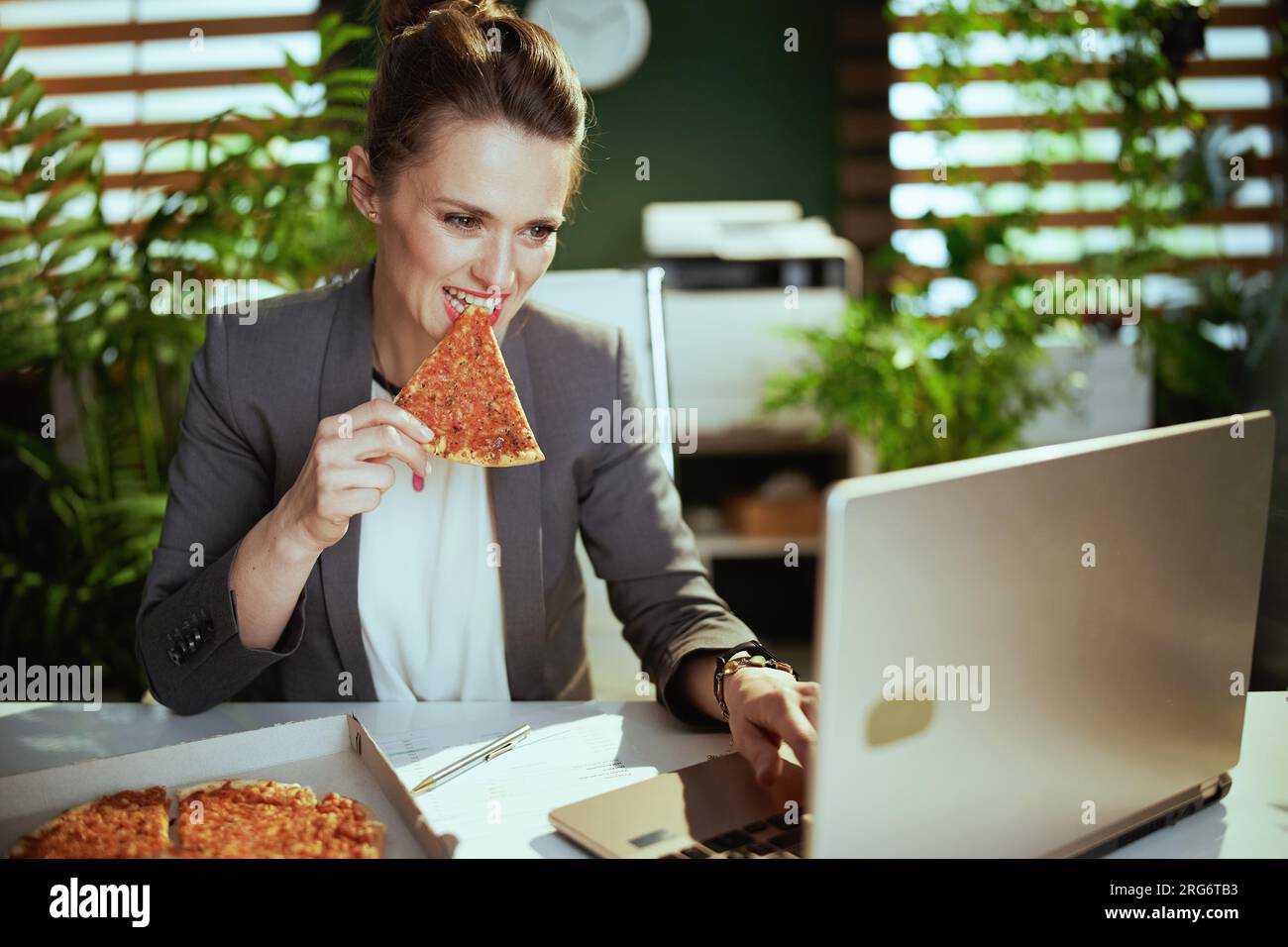 Lieu de travail durable. femme travailleuse moderne dans un costume d'affaires gris dans le bureau vert moderne avec pizza et ordinateur portable. Banque D'Images
