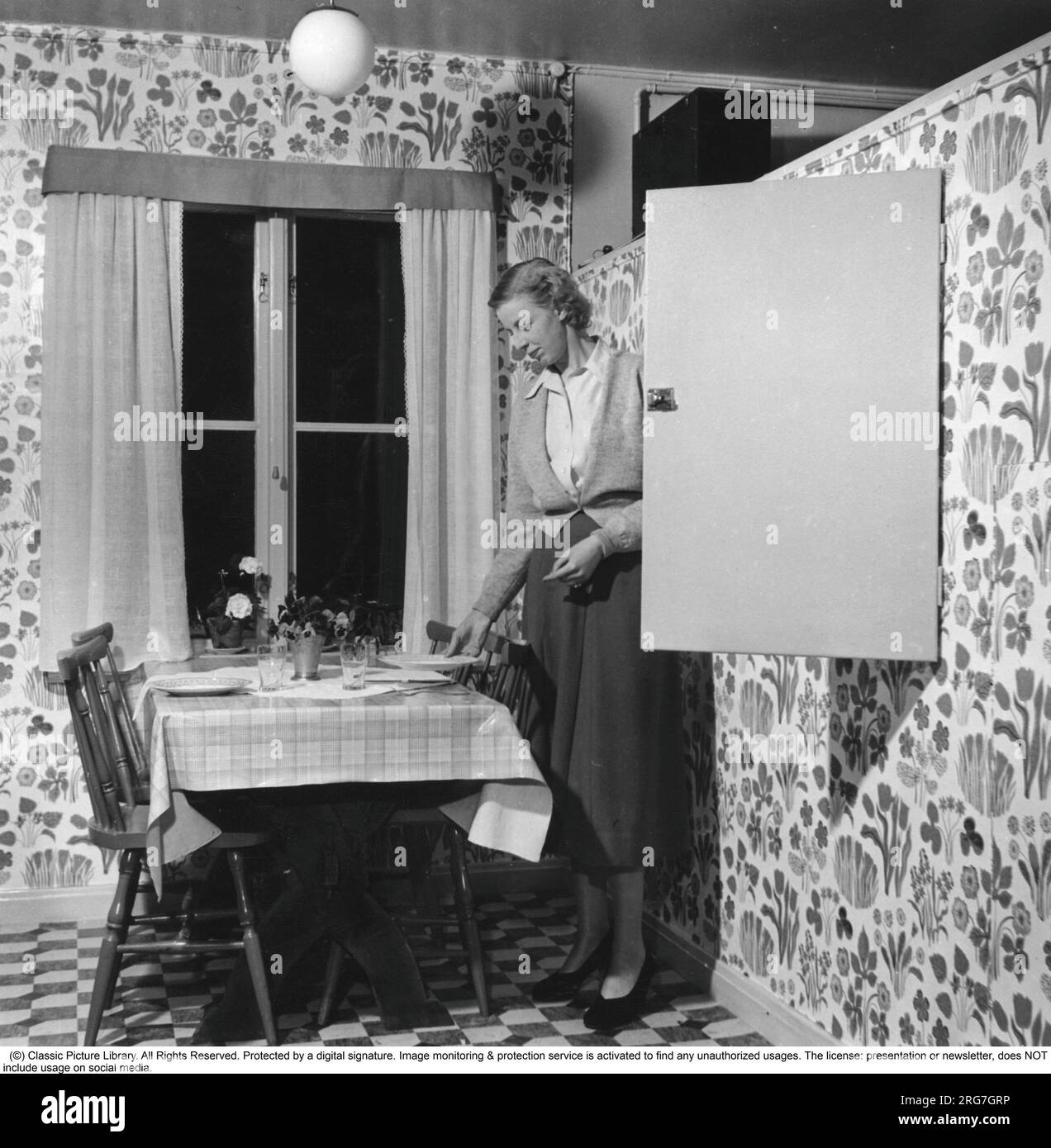 maison des années 1950. Une femme est vue en train de mettre la table pour le dîner et arrange les assiettes et les verres qu'elle prend dans un placard intégré. Suède 1950 Banque D'Images