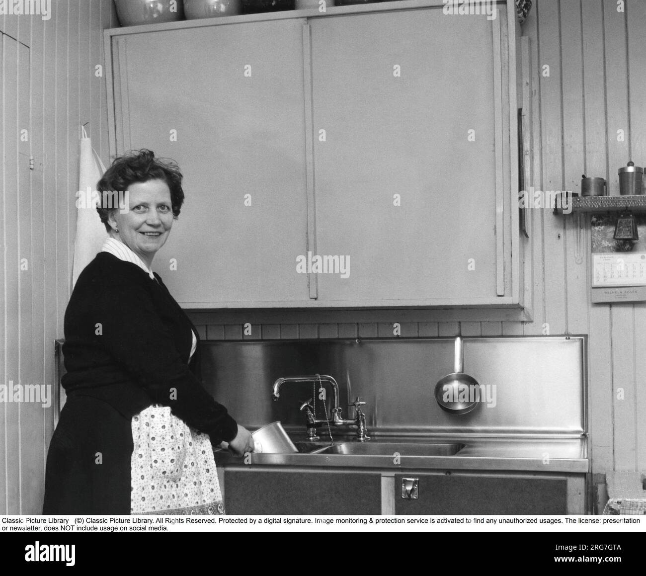 Dans la cuisine 1960s. Intérieur d'une cuisine et une femme nettoyant une casserole dans la cuisine-évier. Suède 1960. Banque D'Images