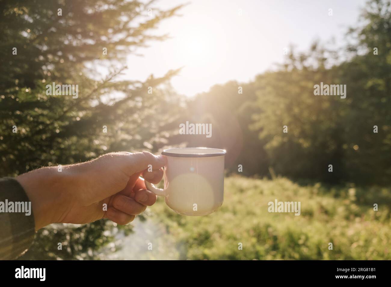 Gros plan de la main masculine tenant le café de tasse sur la forêt de fond et le ciel pendant le lever ou le coucher du soleil chaud. Les touristes masculins boivent du thé chaud et apprécient le paysage Banque D'Images