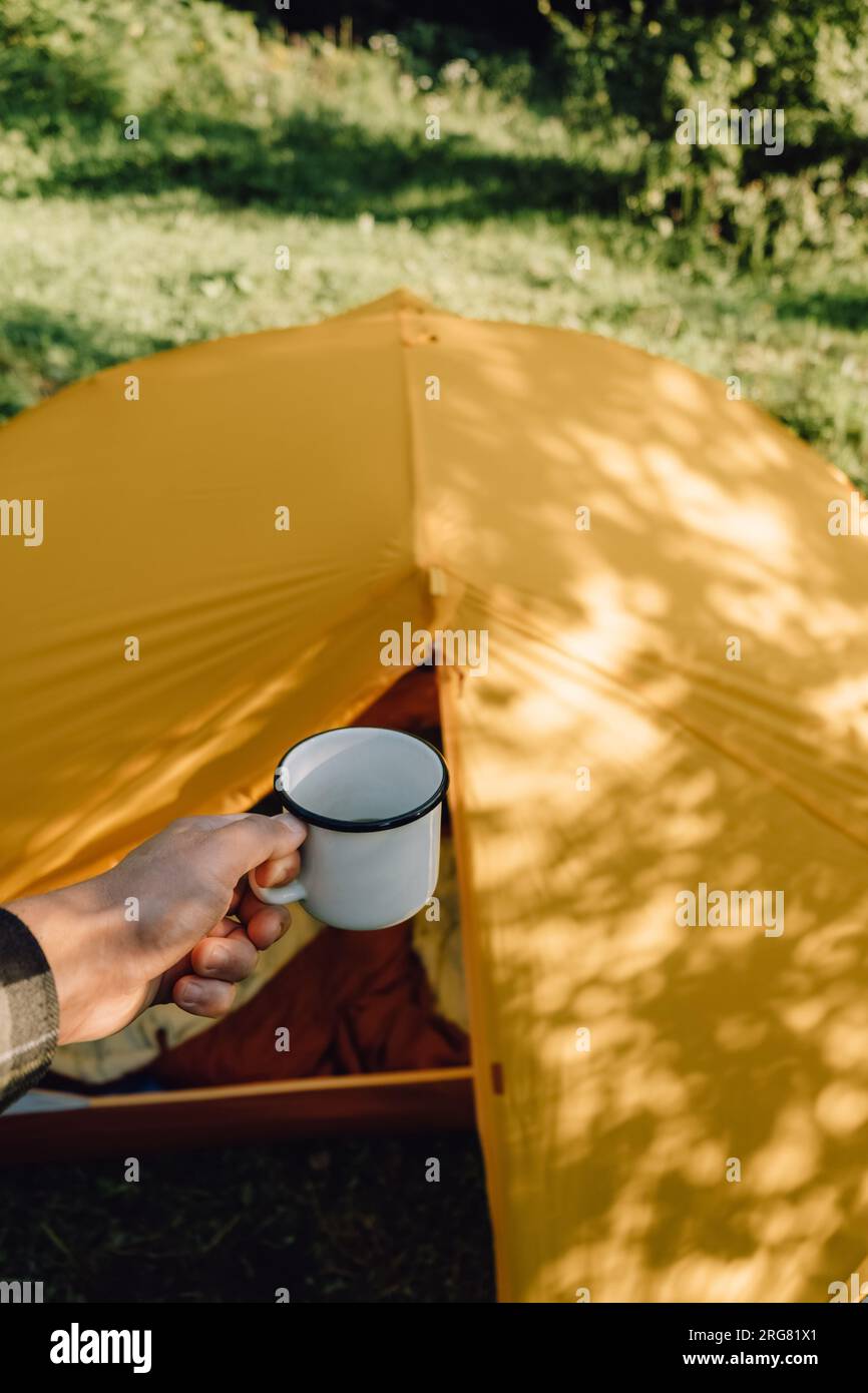 Gros plan d'un jeune homme caucasien tenant une tasse blanche avec du café chaud le matin, profitant de la randonnée. Belles ombres de feuillage d'arbre feuillu reflétées sur le hurlement Banque D'Images