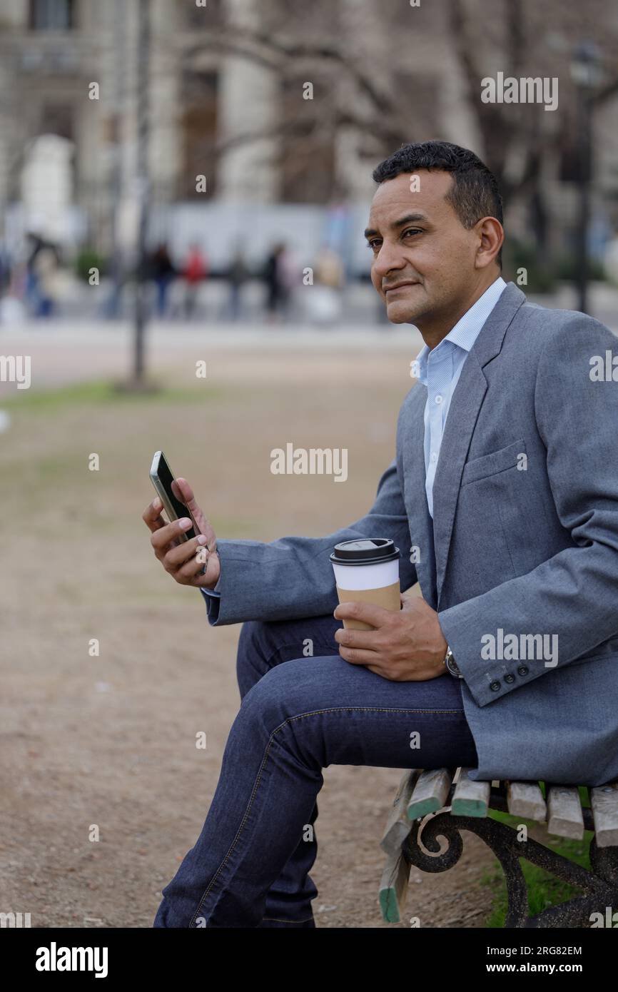 Portrait d'homme latino en veste avec une tasse de café jetable assis sur un banc dans un parc public. Banque D'Images