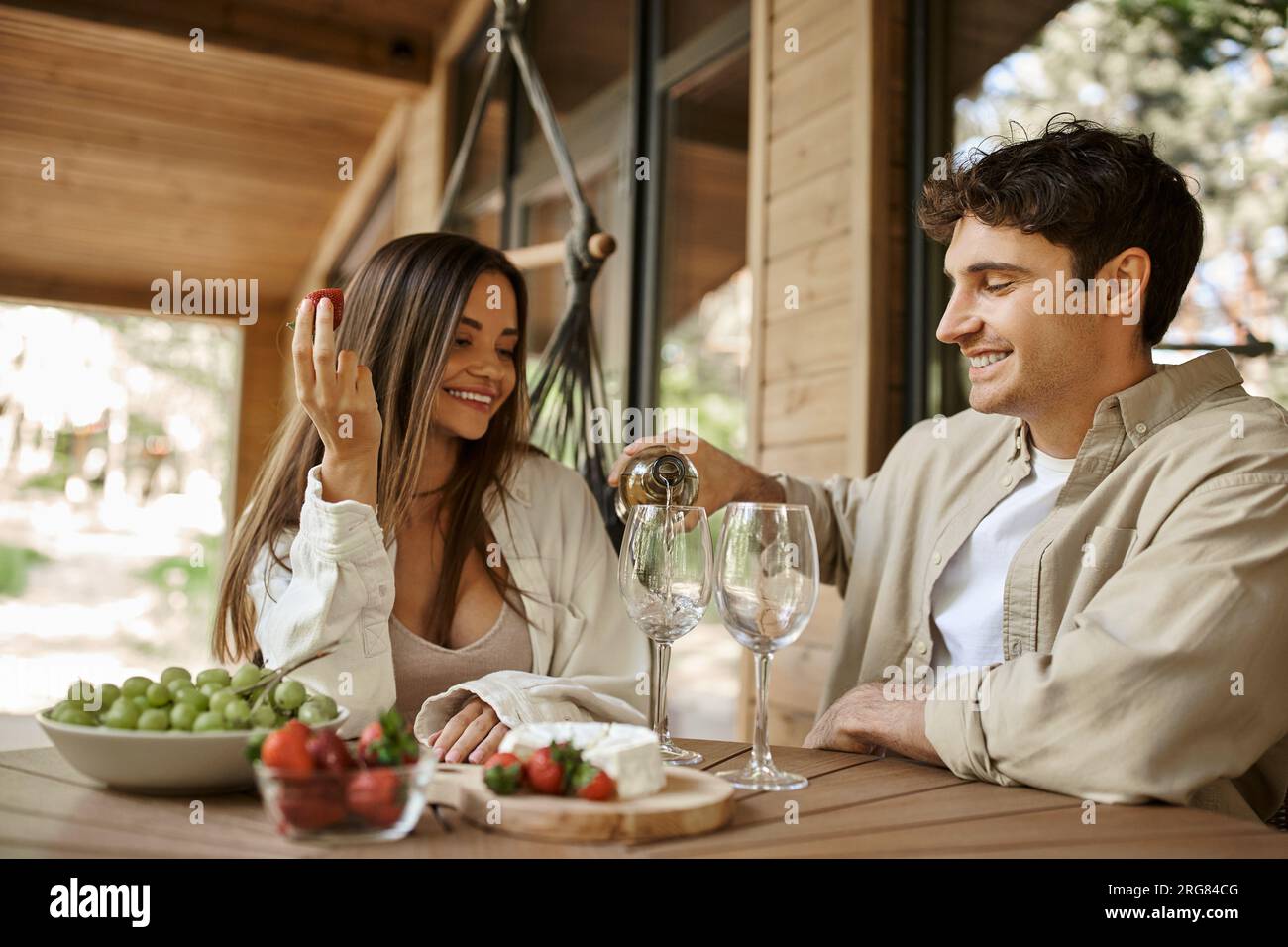 Homme souriant versant du vin près de petite amie avec fraise sur la terrasse de la maison de vacances à l'arrière-plan Banque D'Images