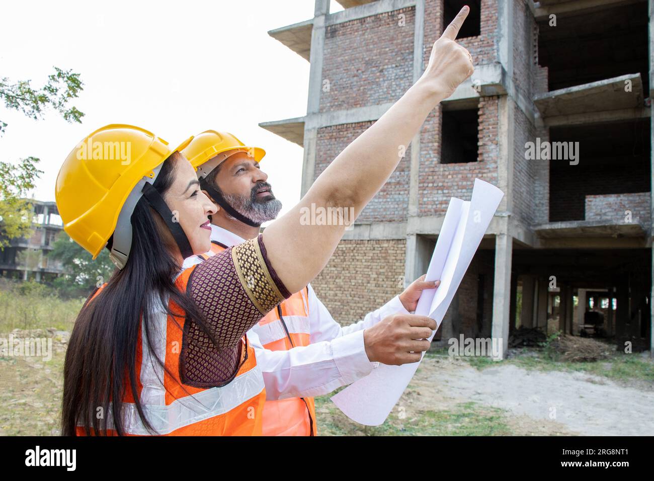 Deux ingénieurs civils indiens hommes et femmes ou un architecte portant casque et gilet tenant le plan de paperasse sur le chantier de construction discutant du vrai estat Banque D'Images