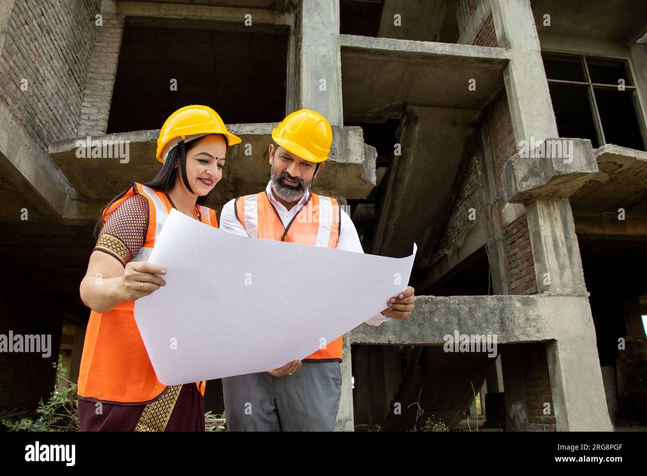 Deux ingénieurs civils indiens hommes et femmes ou un architecte portant casque et gilet tenant le plan de paperasse sur le chantier de construction discutant du vrai estat Banque D'Images