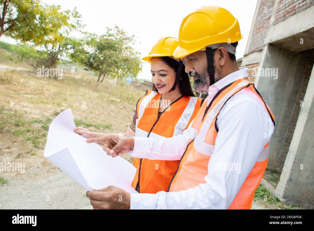Deux ingénieurs civils indiens hommes et femmes ou un architecte portant casque et gilet tenant le plan de paperasse sur le chantier de construction discutant du vrai estat Banque D'Images