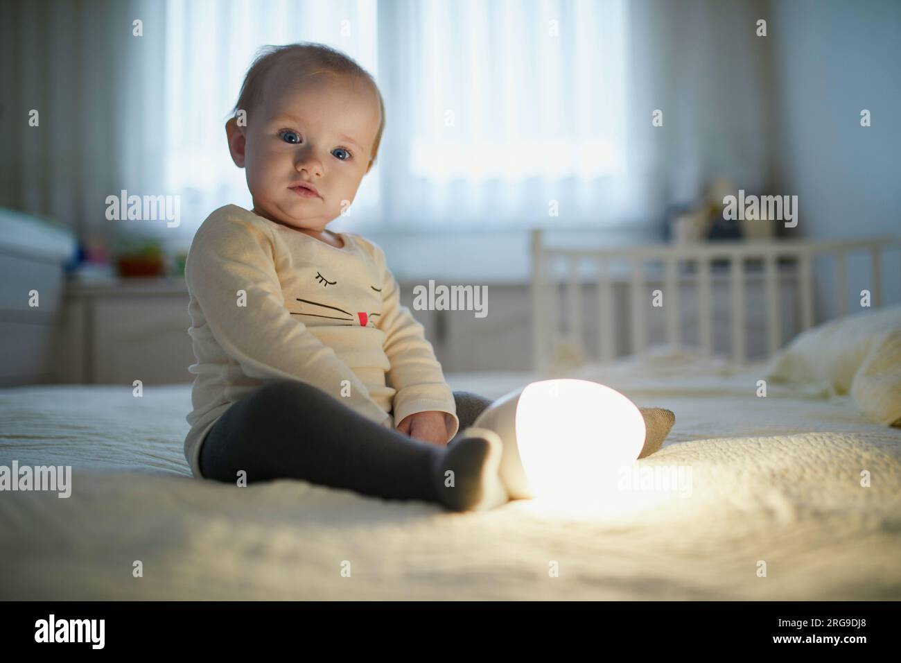 Adorable petite fille jouant avec la lampe de chevet dans la pépinière. Enfant heureux assis sur le lit avec veilleuse. Petit enfant à la maison le soir avant de dormir Banque D'Images