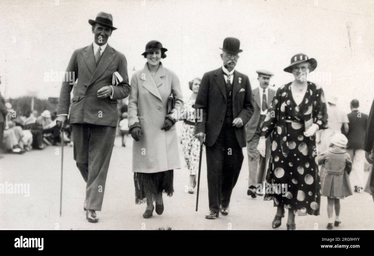 Un couple britannique chic et ultra-élégant de 1930s vous promènerez le long du front de mer d'une station balnéaire avec les parents de la jeune femme, la mère portant une robe à imprimé assez remarquable, qui est ornée de grands fruits ou de planètes !!! Banque D'Images
