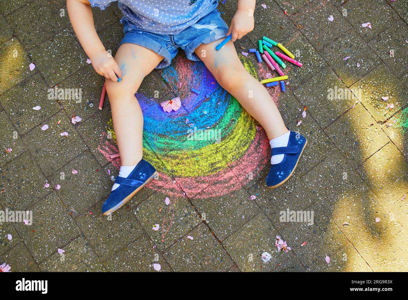 Petite fille dessinant arc-en-ciel avec des craies colorées sur l'asphalte. Enfant s'amusant à peindre ses jambes. Activités de plein air et jeux créatifs pour les petits enfants Banque D'Images